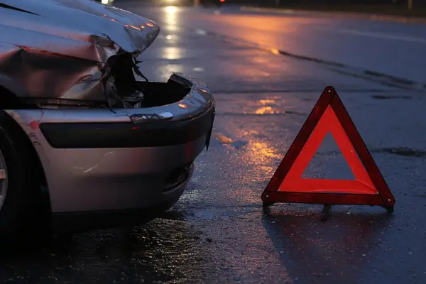 Illustration: the aftermath of a collision — a damaged bumper and a hazard triangle on wet asphalt.